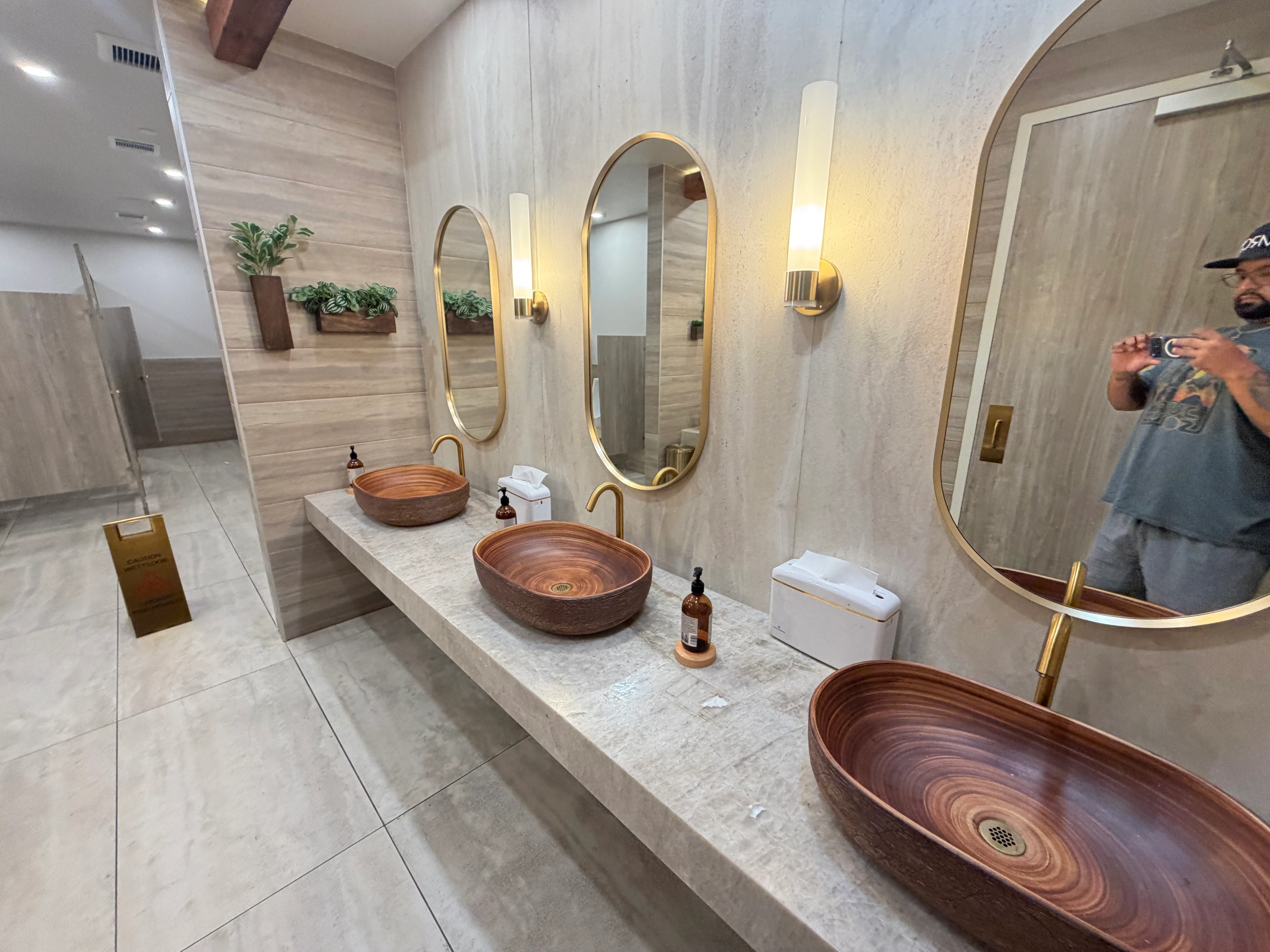 Luxury bathroom with marble countertop, three wood vessel sinks, brass mirrors, and tile walls