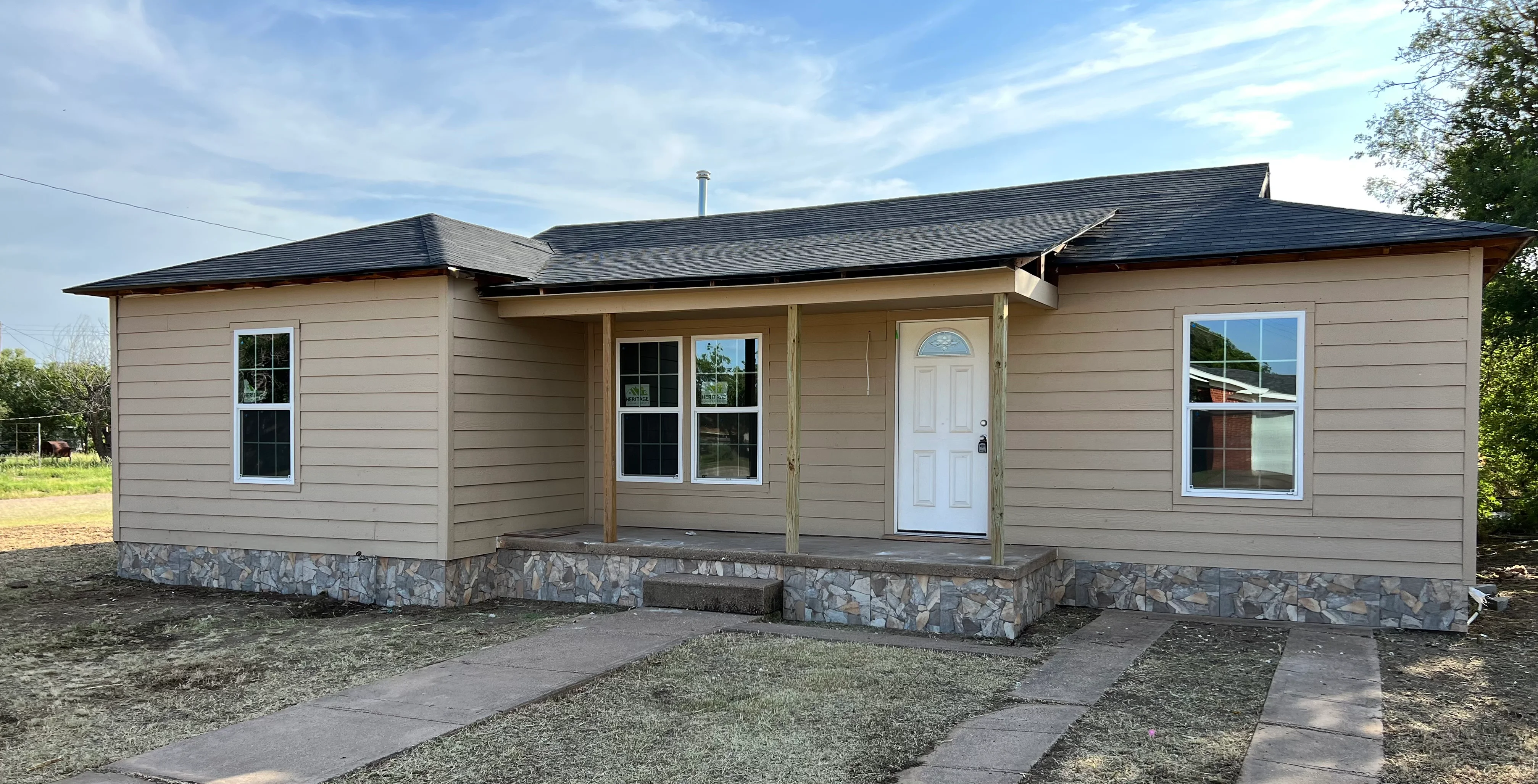 Modern home with tan siding, dark roof, stone foundation, and covered porch entry