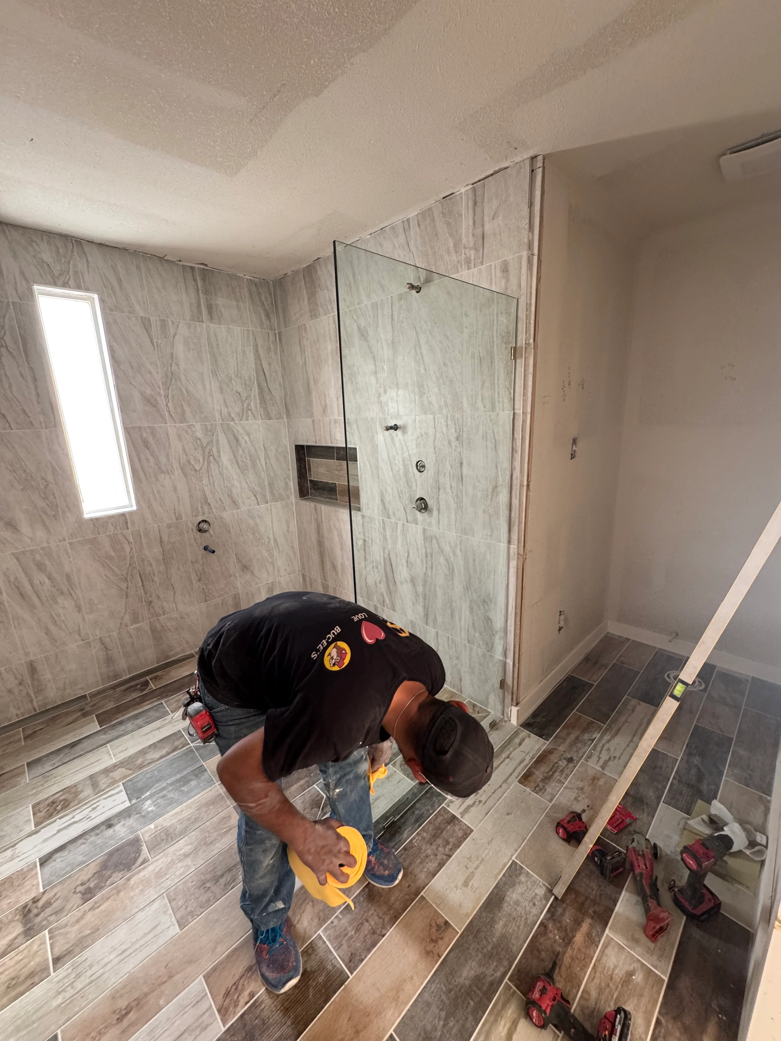 Newly remodeled shower with gray wood-grain tile, frameless glass door, and dual showerheads
