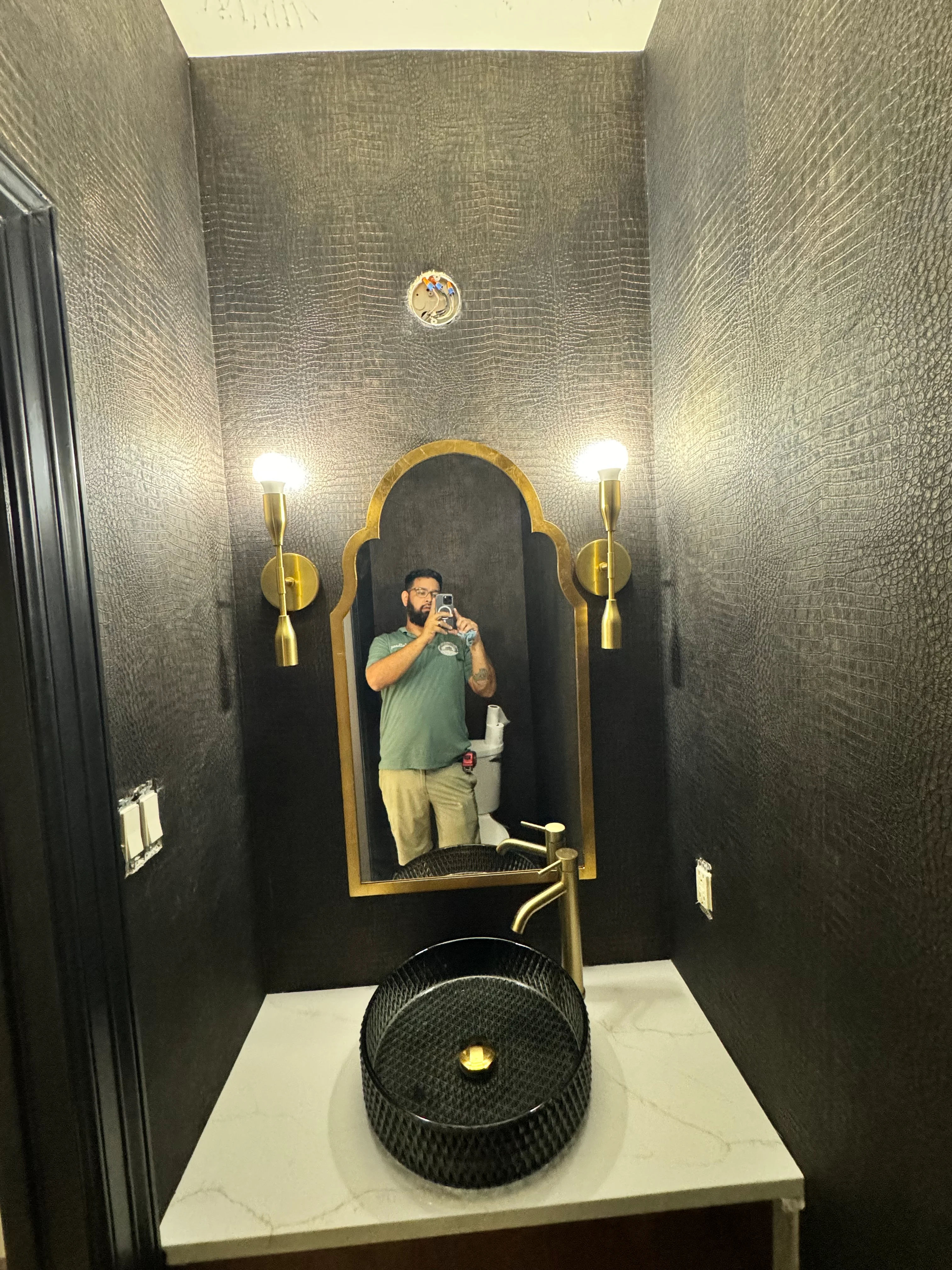 Modern powder room with gray textured tile walls, black vessel sink, brass fixtures, and arched mirror
