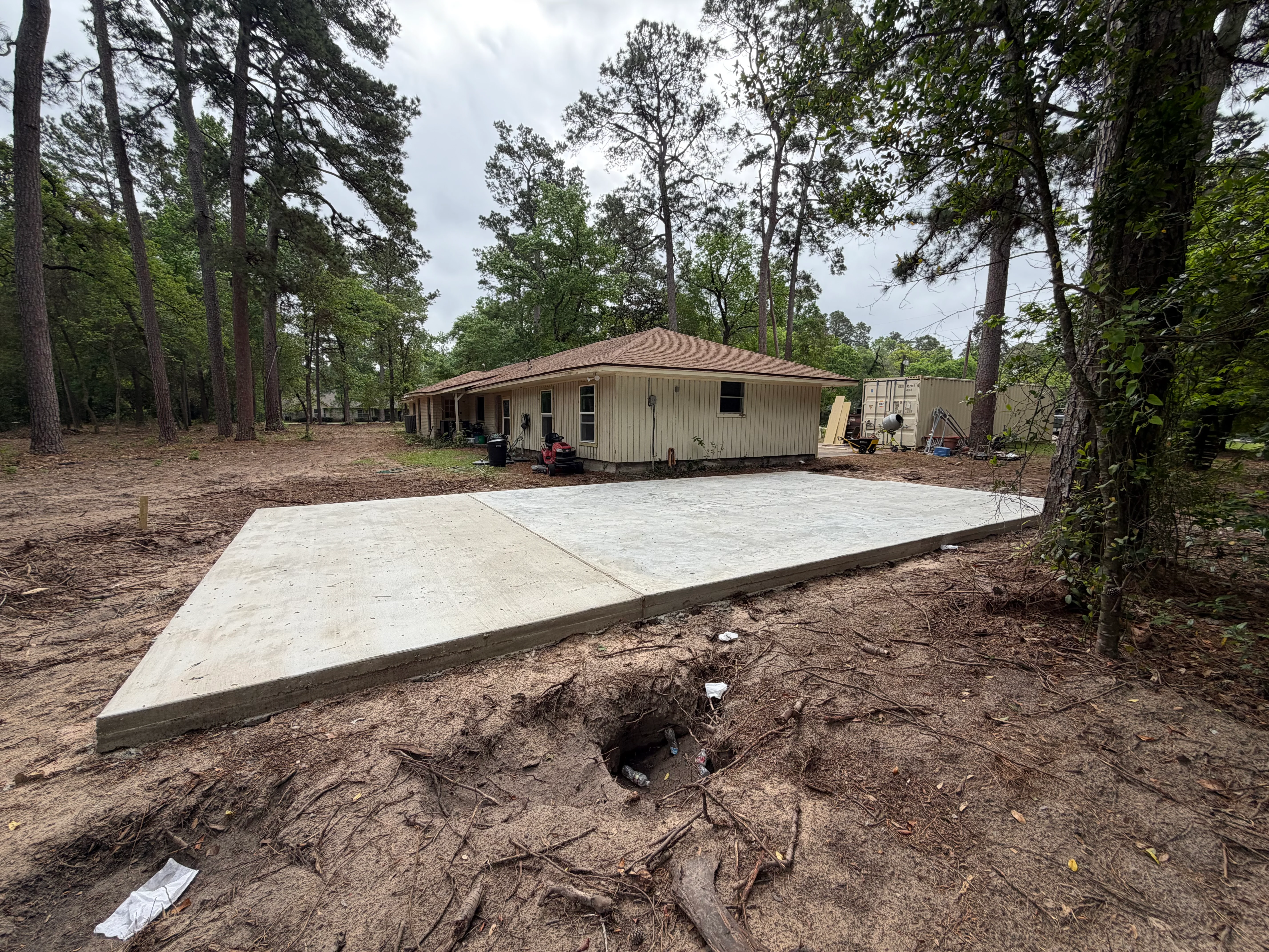 Newly poured concrete foundation pad in front of metal-frame residential structure surrounded by tall pine trees
