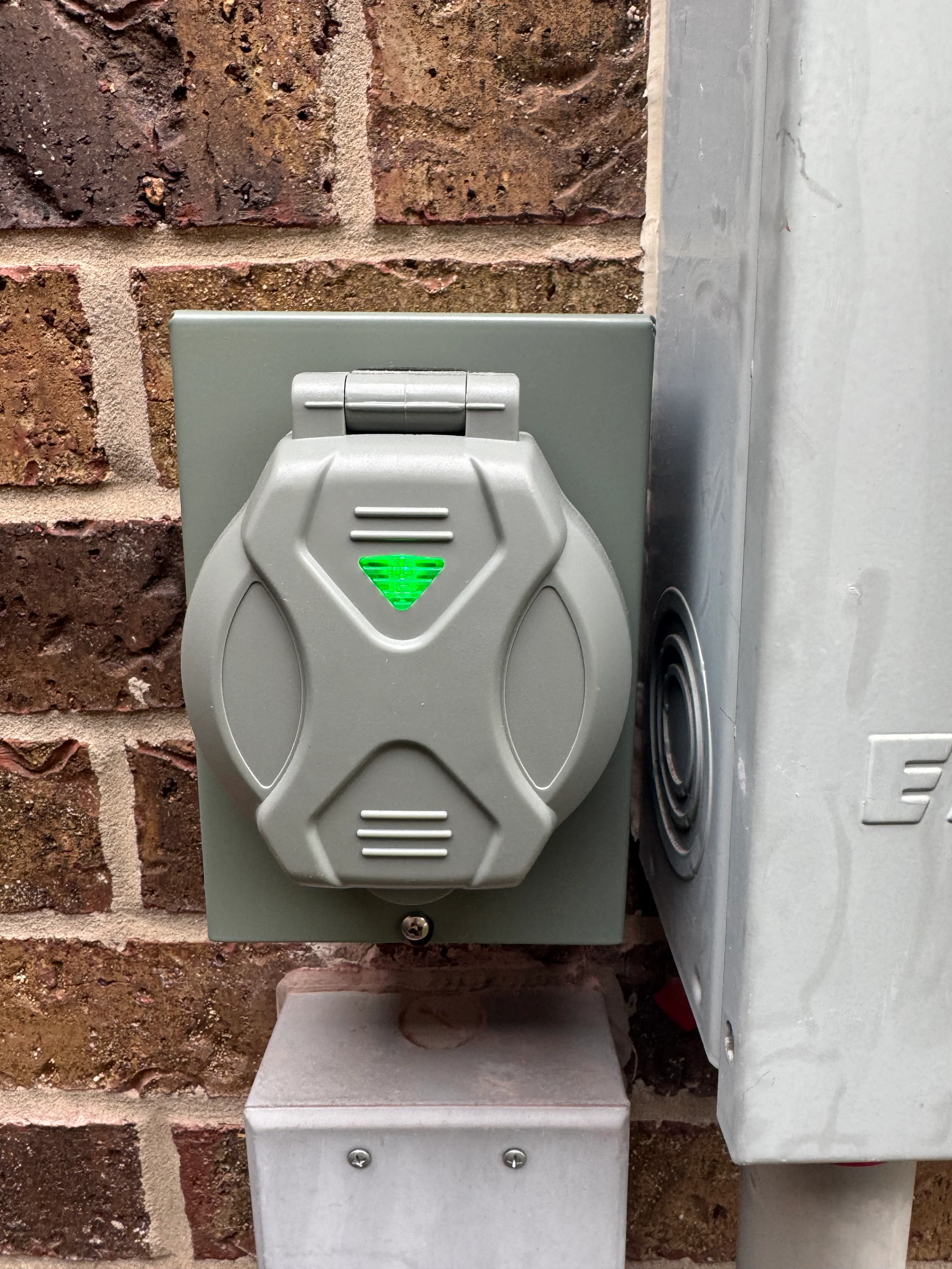 Gray weatherproof electrical outlet with green indicator mounted on brick exterior wall