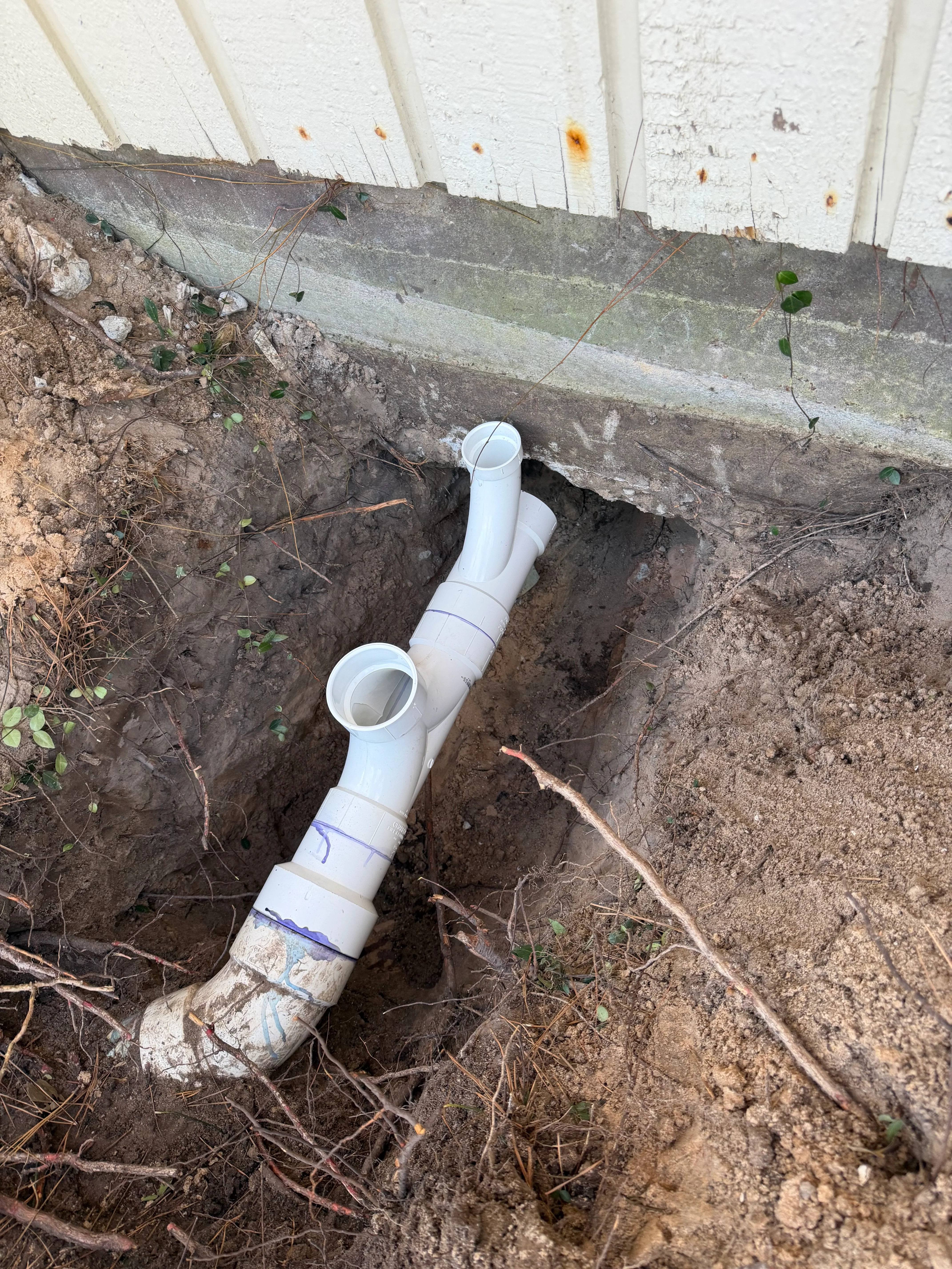 White PVC drainage pipes with fittings installed in excavated trench with soil and foundation visible