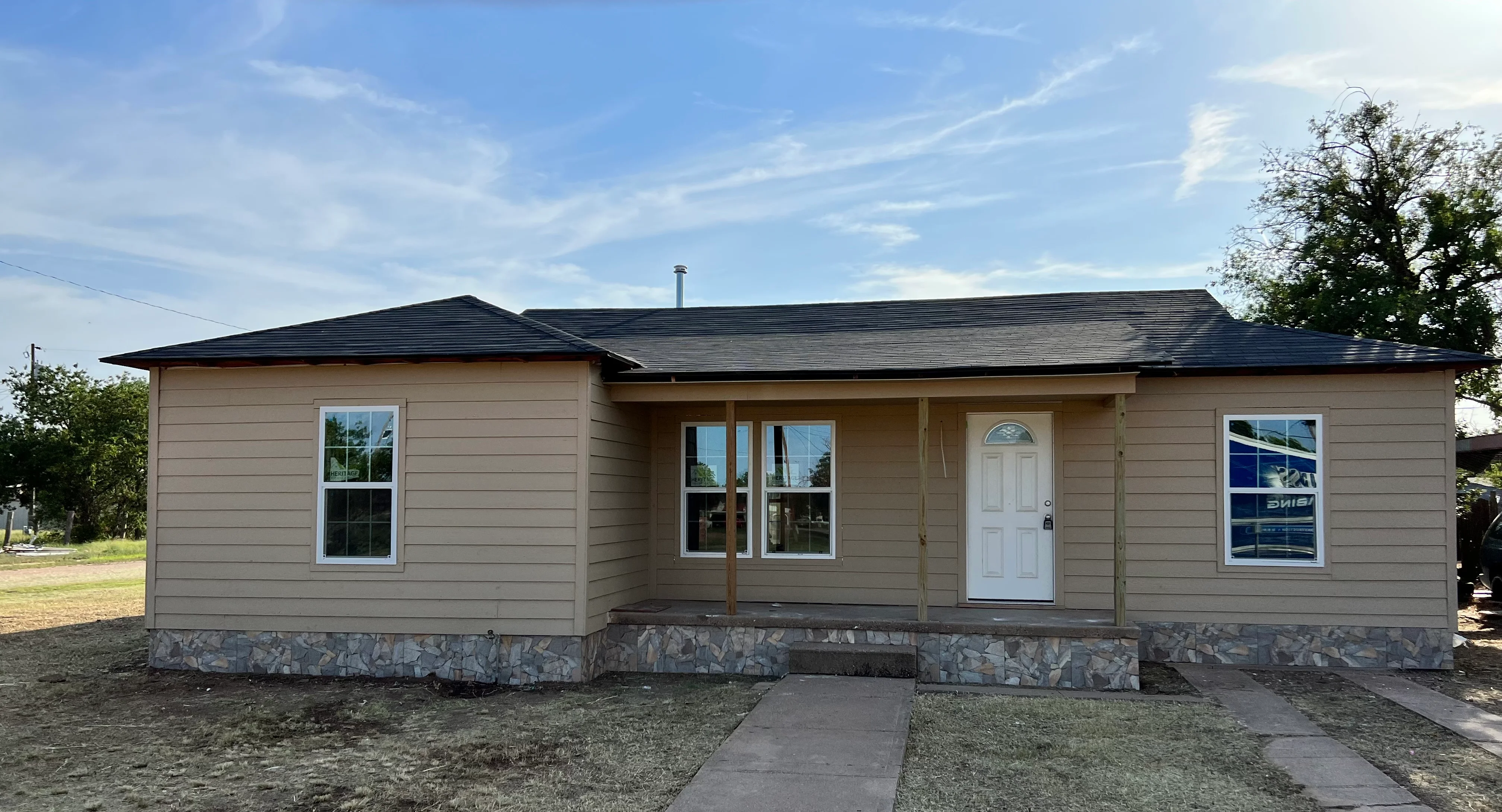 Modern ranch home with tan siding, dark roof, stone foundation, and covered porch entry