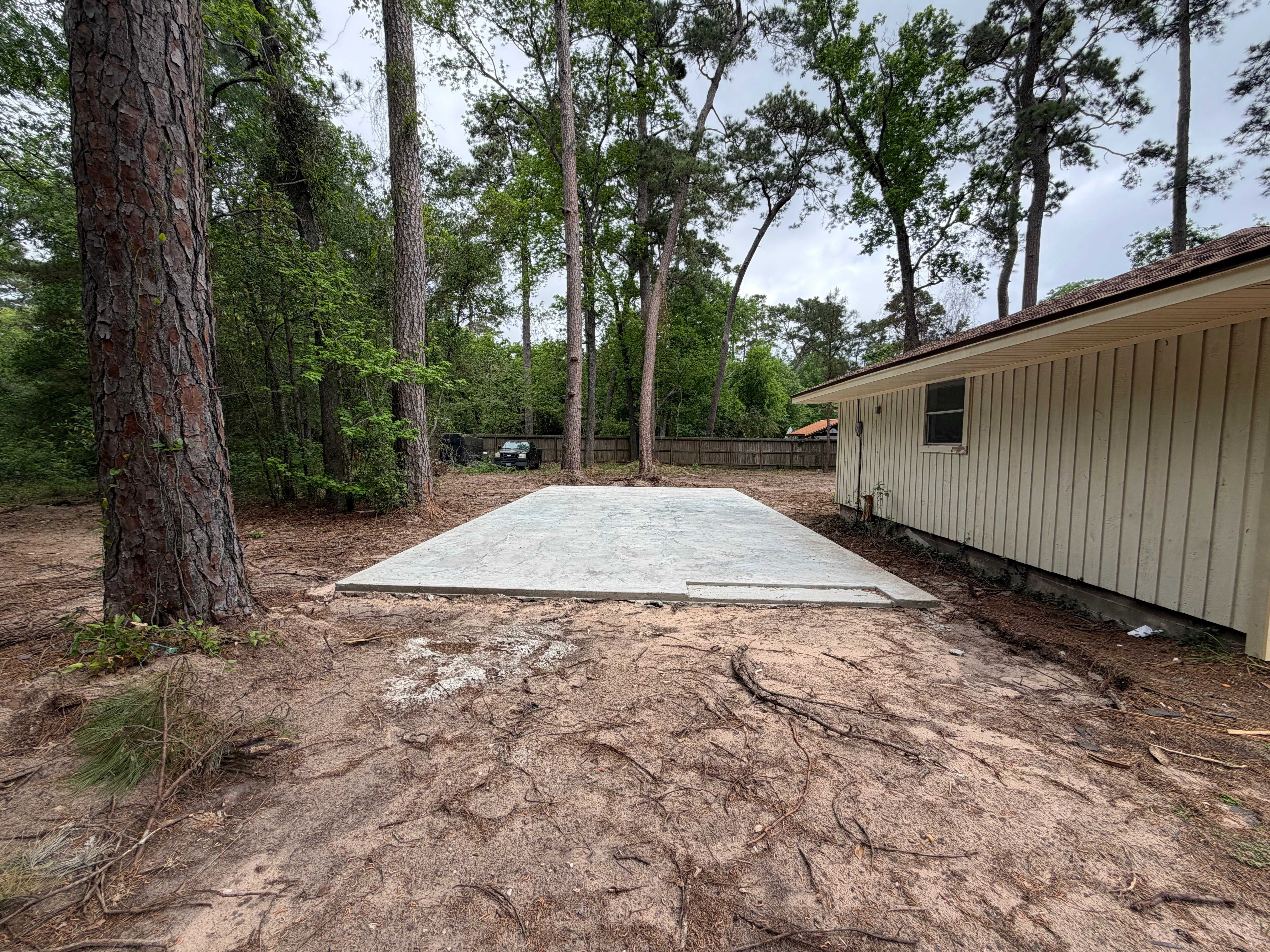 Newly poured concrete foundation pad in wooded backyard with mature pine trees