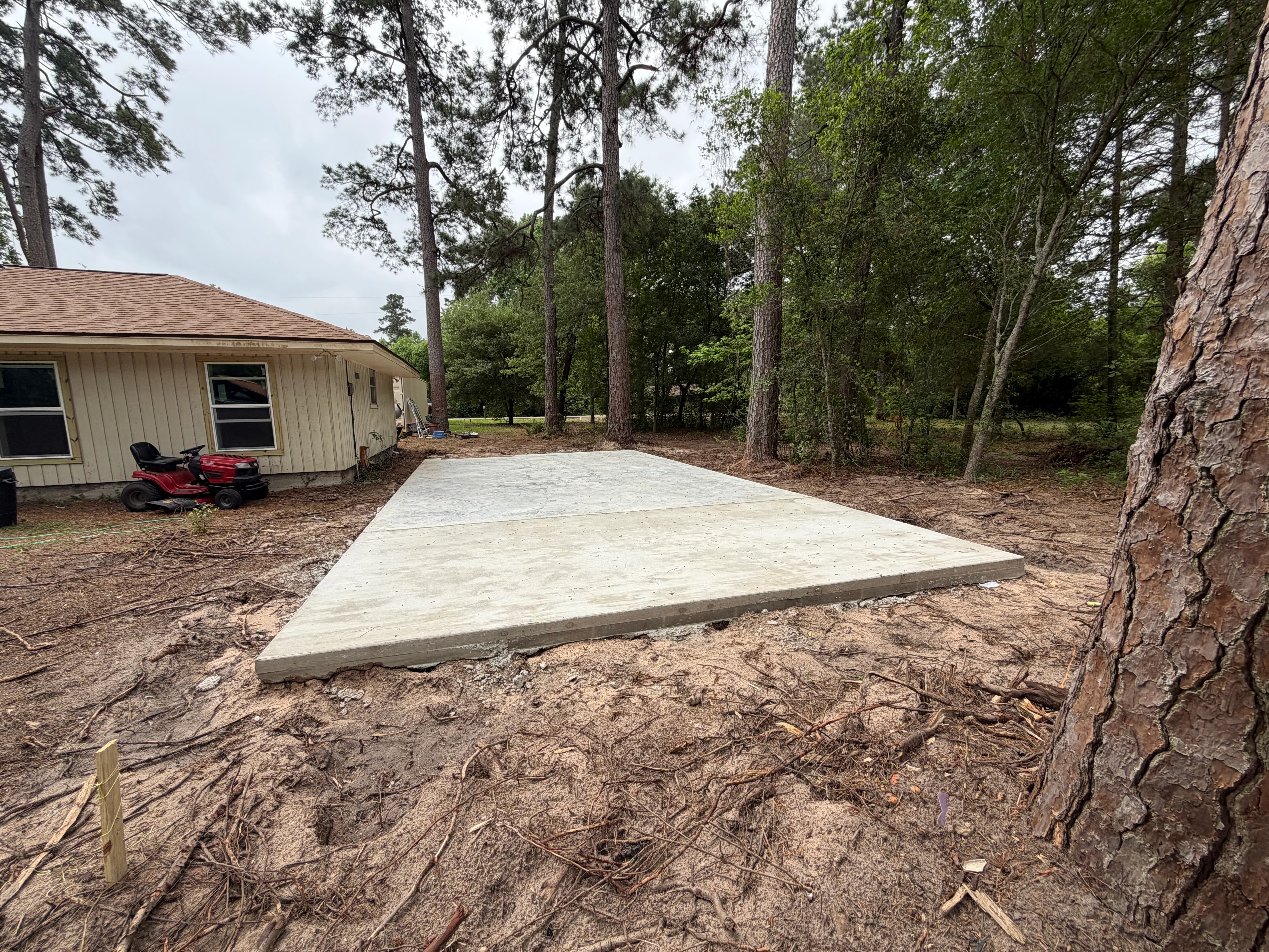 Newly poured concrete pad next to residential building surrounded by tall pine trees