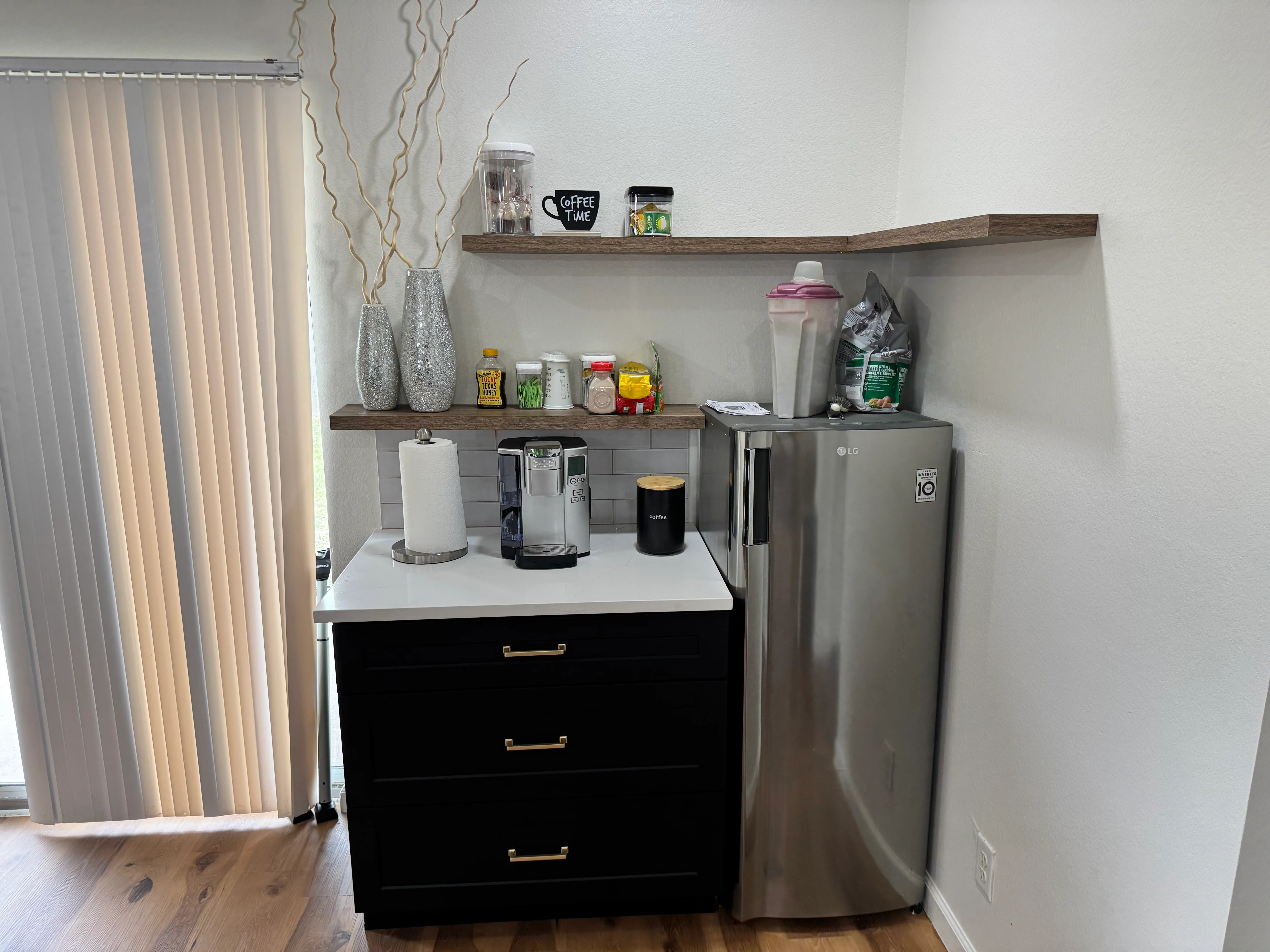 Modern kitchen beverage station with black cabinetry, floating shelves, and stainless steel refrigerator
