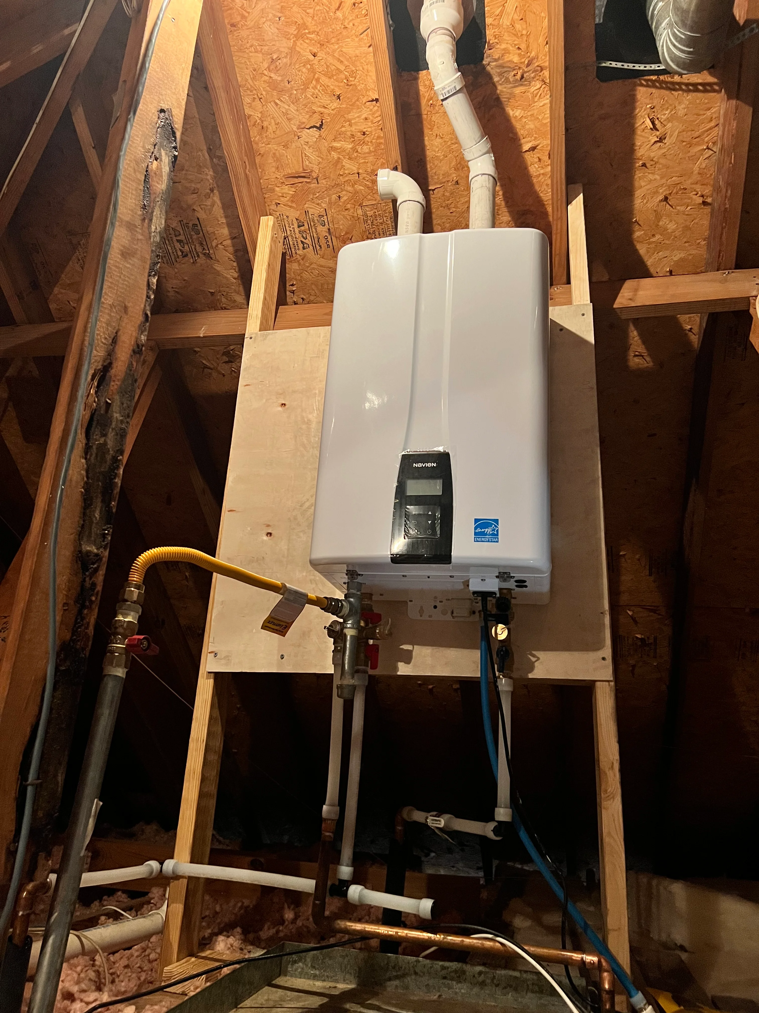 Tankless water heater installation with white unit mounted on wooden frame in attic space