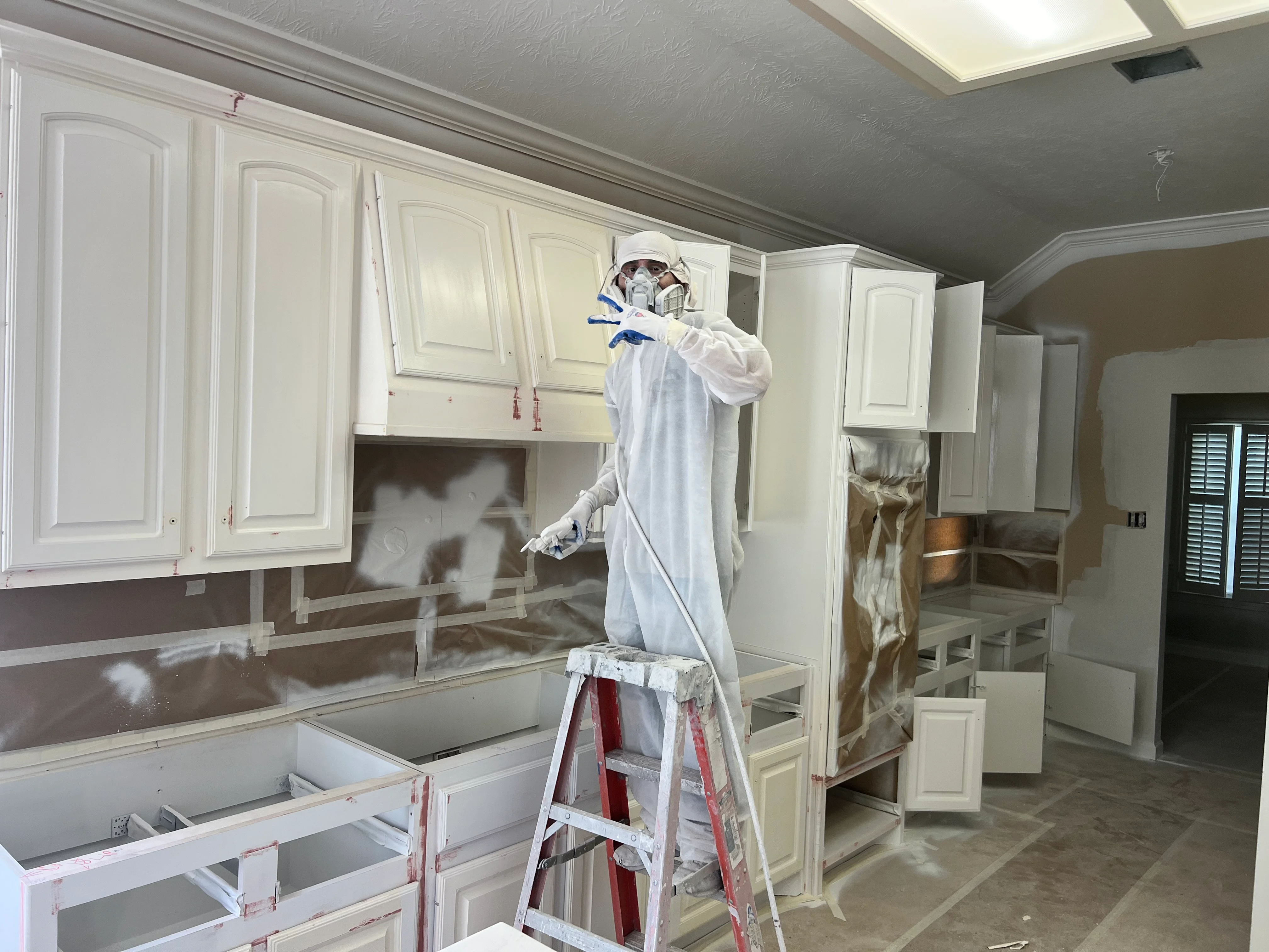 Professional painter in protective gear refinishing cream-colored kitchen cabinets with spray equipment