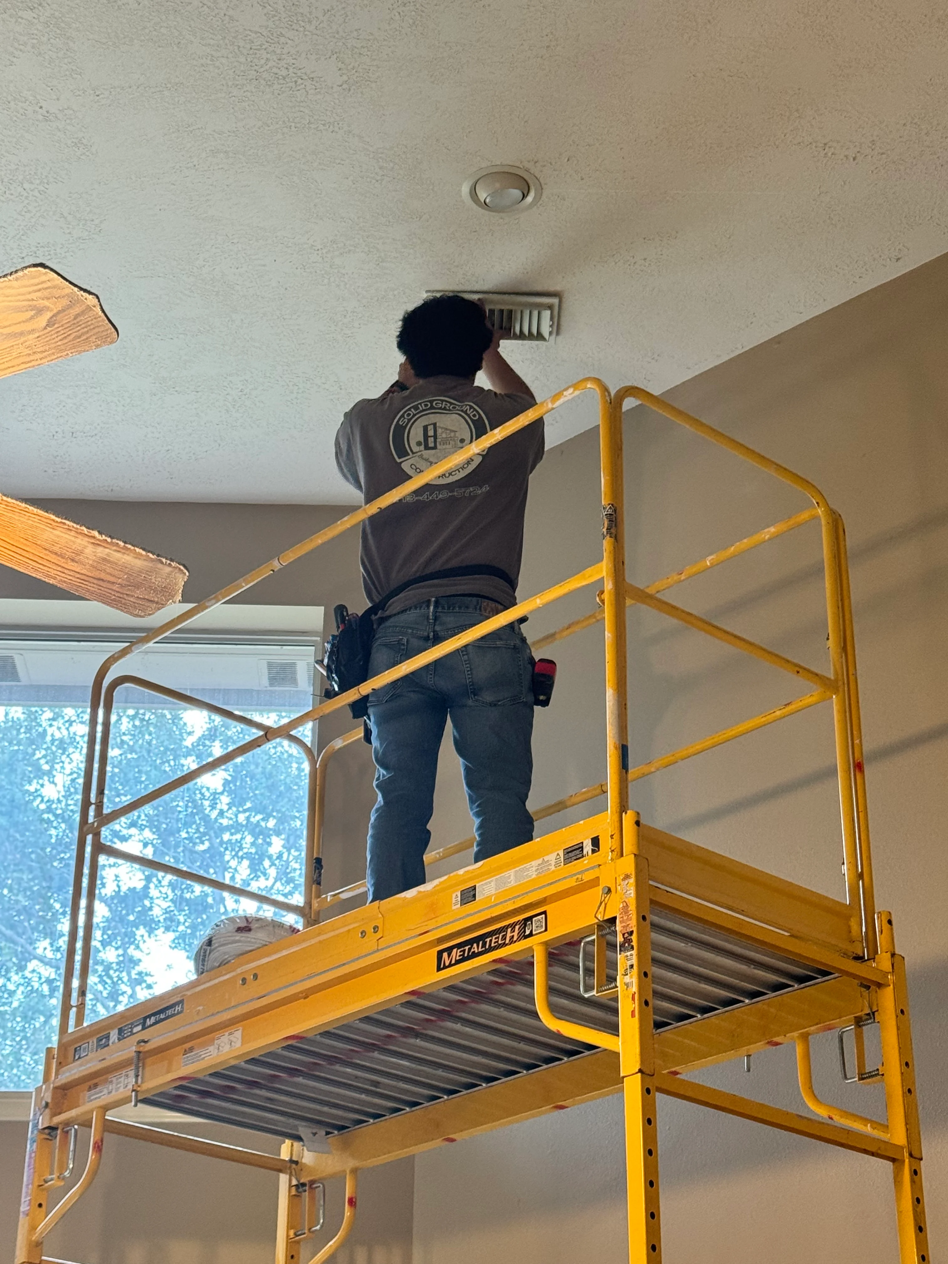 Worker on yellow scaffolding installing recessed light fixture in textured ceiling