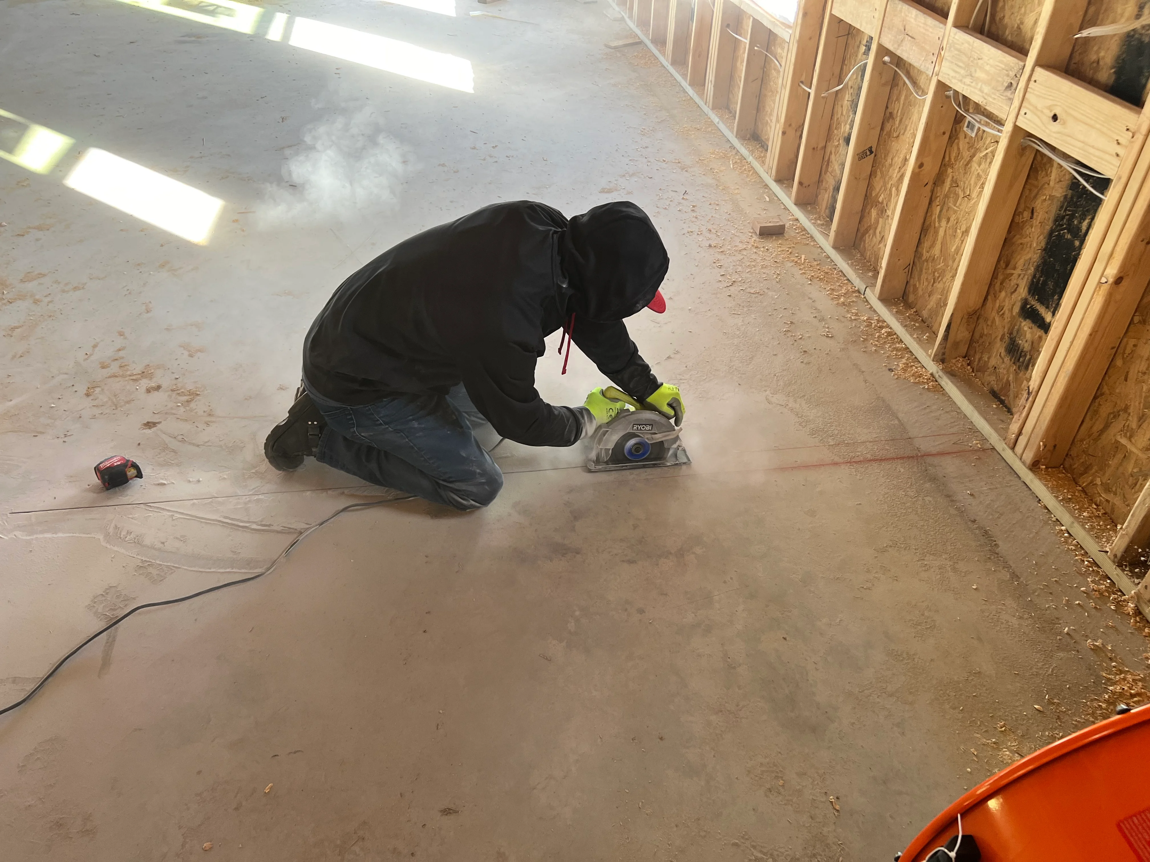 Worker using circular saw to cut concrete floor in residential space with wooden stalls visible