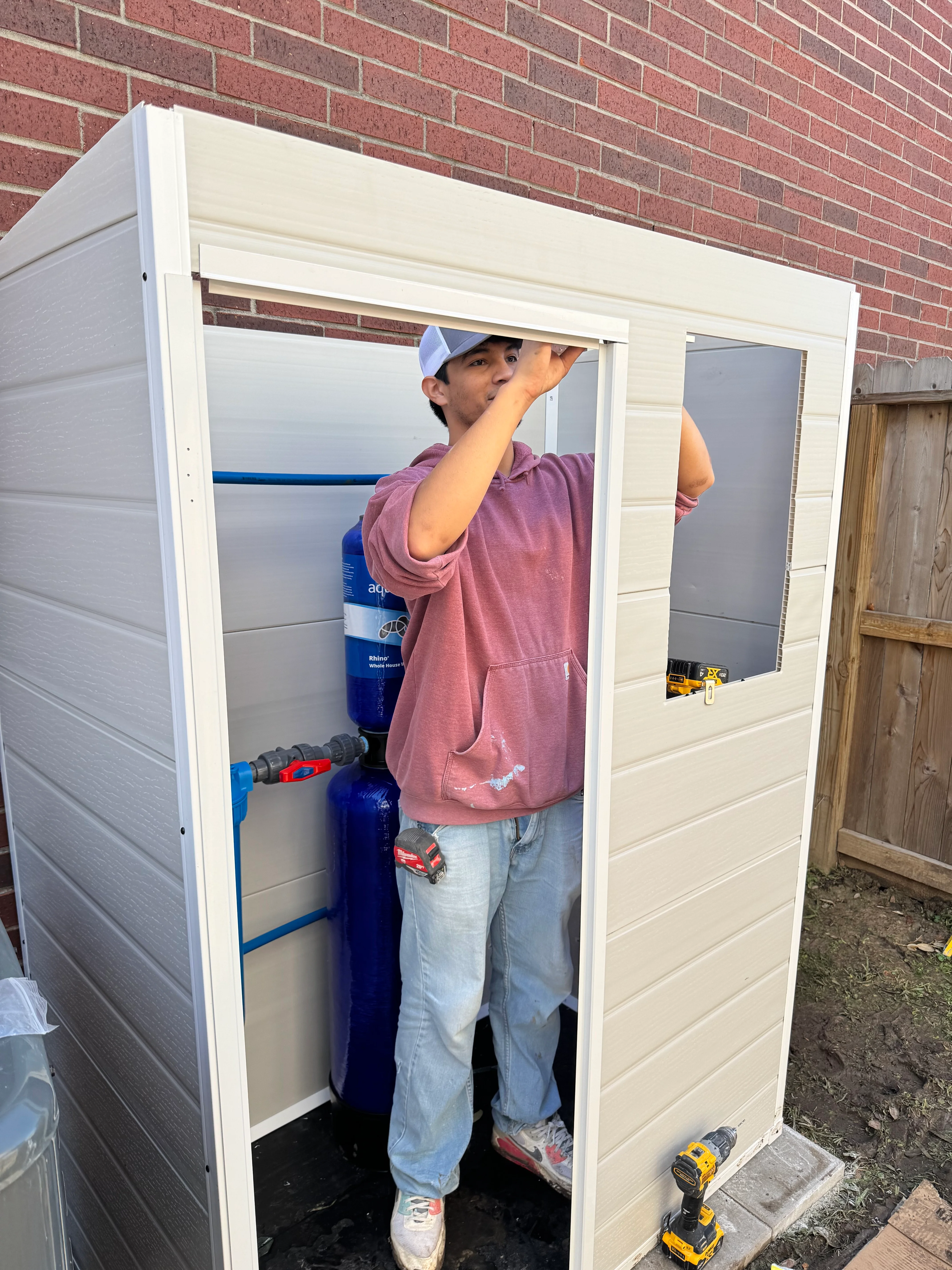 Technician installing cream vinyl utility shed with windows against brick home