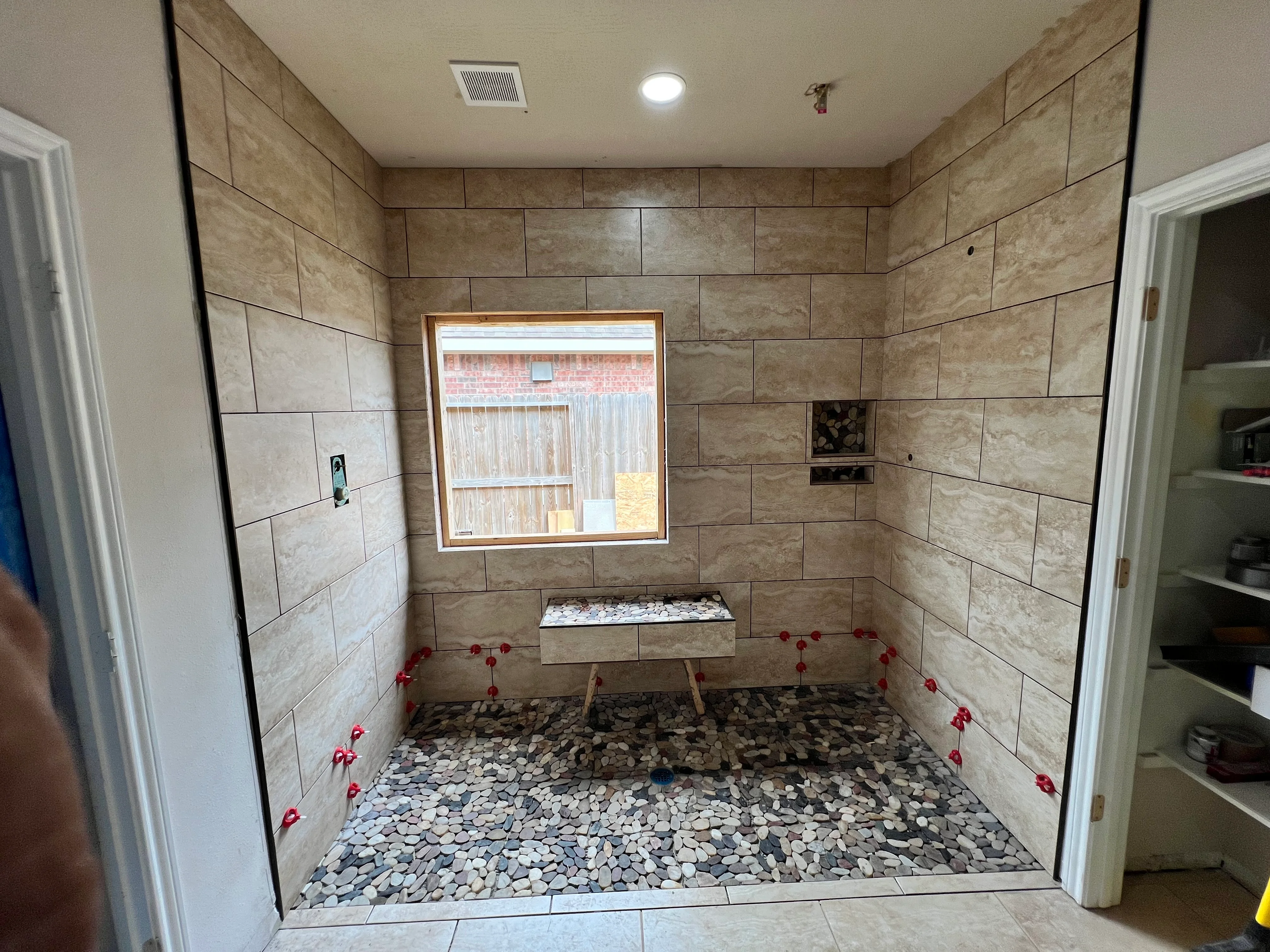 Newly tiled shower enclosure with beige porcelain tiles, pebble floor, and wooden-framed window
