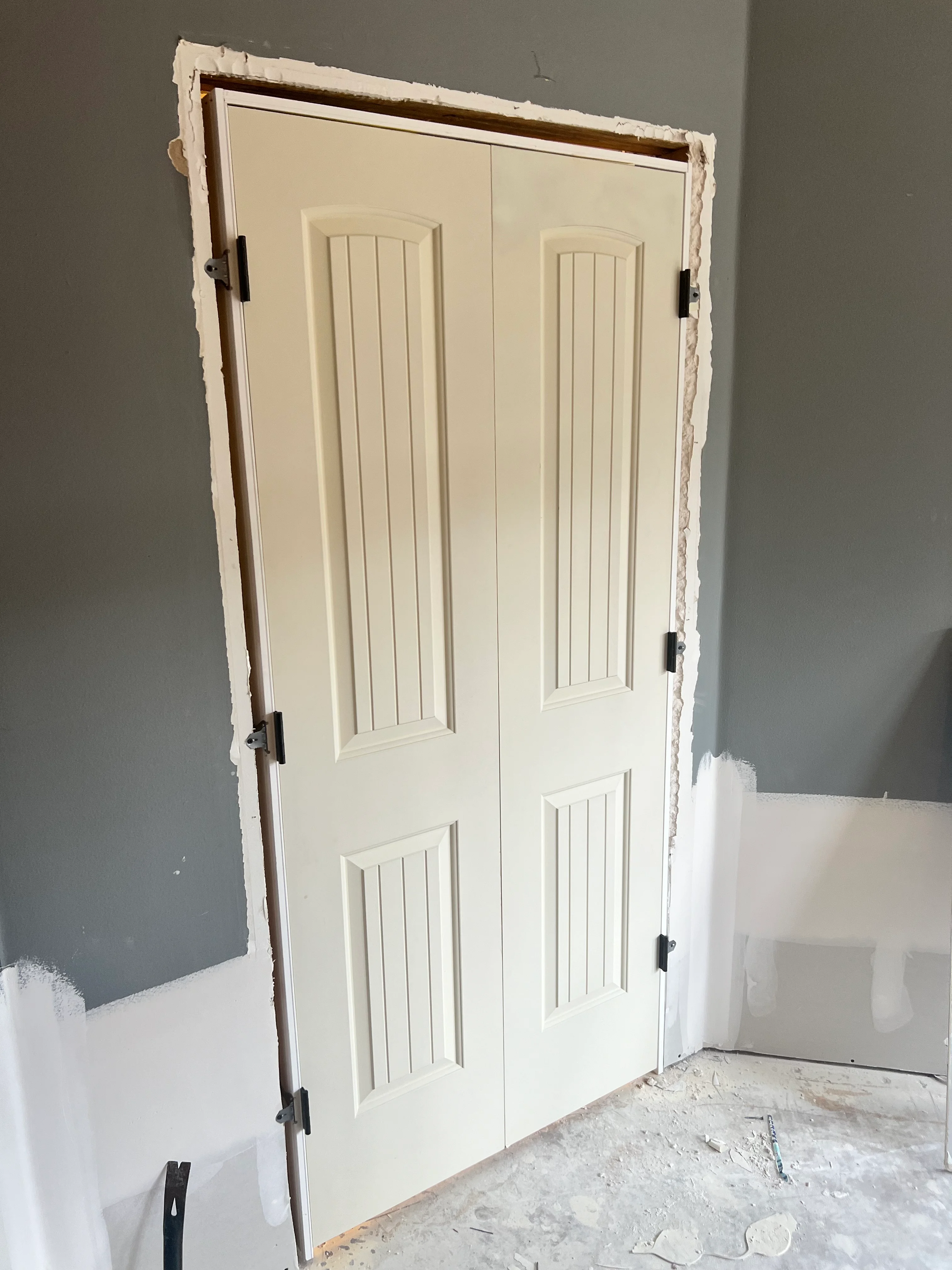 Cream double doors with vertical panels and black hinges installed in drywall opening