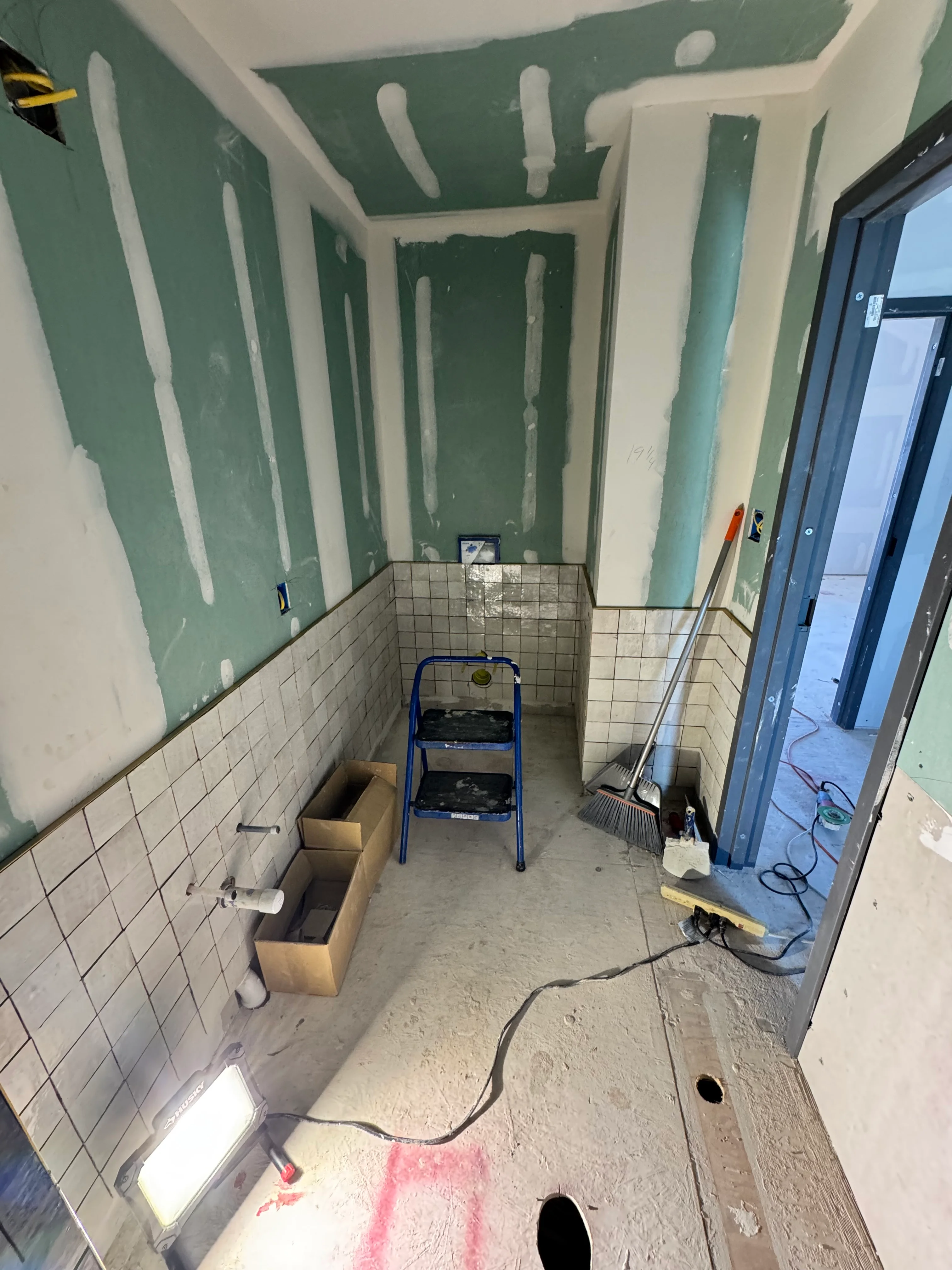 Bathroom under renovation with green drywall compound, white tile wainscot, and plumbing preparations